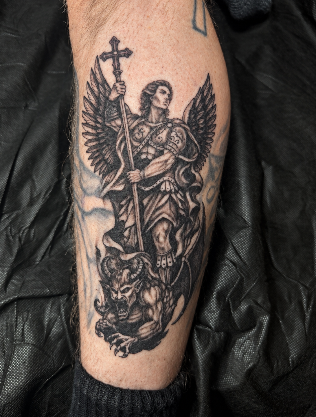 A black-and-gray winged warrior holding a cross-topped weapon above a fallen creature. Tattoo work by Joseph Rojas.